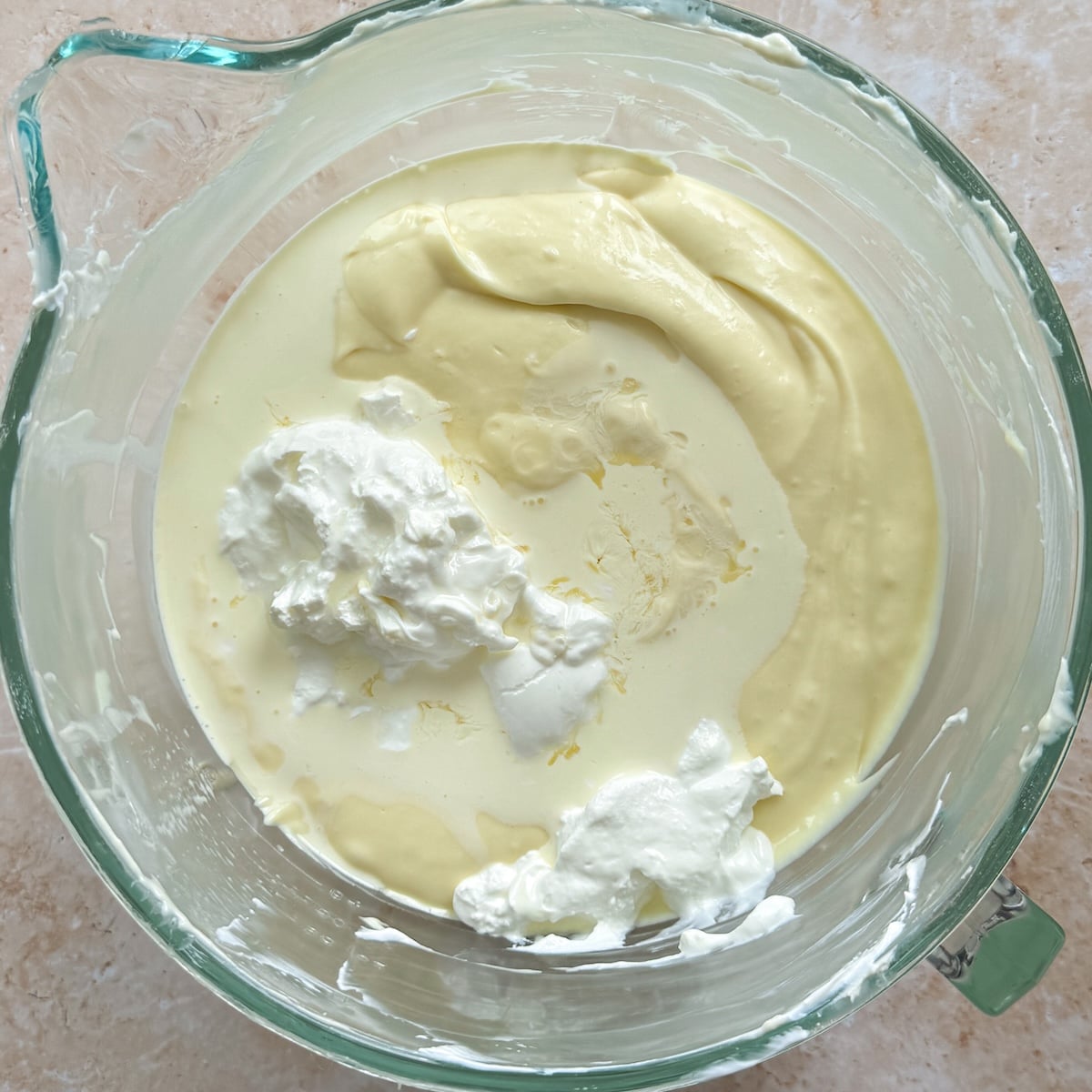 Glass bowl with thick, creamy yellow cheesecake mixture and dollops of white sour cream and heavy cream, viewed from above, on a light countertop.