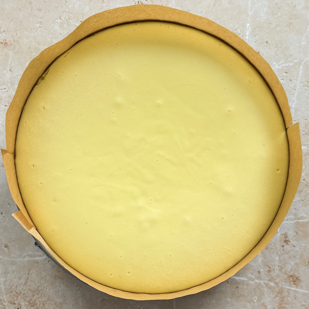 A classic cheesecake with a smooth, creamy yellow surface sits in a baking pan lined with parchment paper on a light marble surface. The plain, round cheesecake showcases its flawless texture and inviting simplicity.