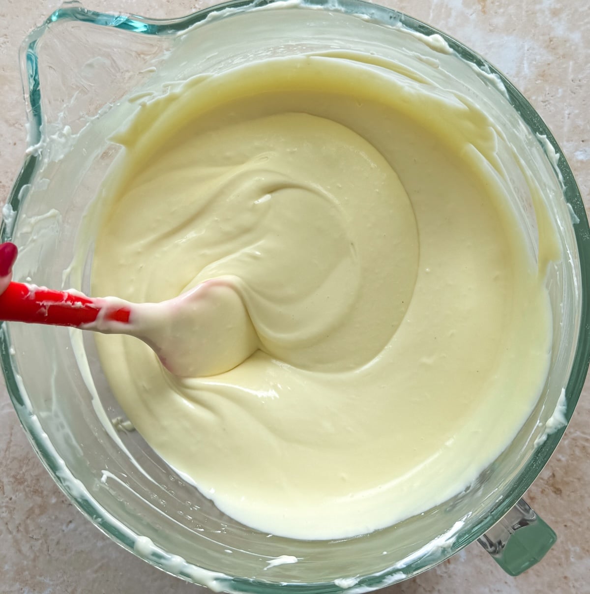 A glass mixing bowl filled with smooth, creamy yellow cheesecake batter. A red and white spatula rests inside, partially buried in the thick mixture. The bowl sits on a light-colored countertop.