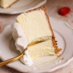 a close up picture of a slice of cheesecake placed sideways on a small plate with a fork taking out a bite. the cheesecake has a very creamy, silky texture, a crispy golden crust, and whipped cream on top
