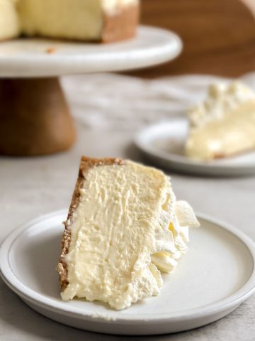 Close up of a slice of cheesecake placed sideways on a small plate with a thin crust and very creamy interior. Another slice and full cheesecake in the background.