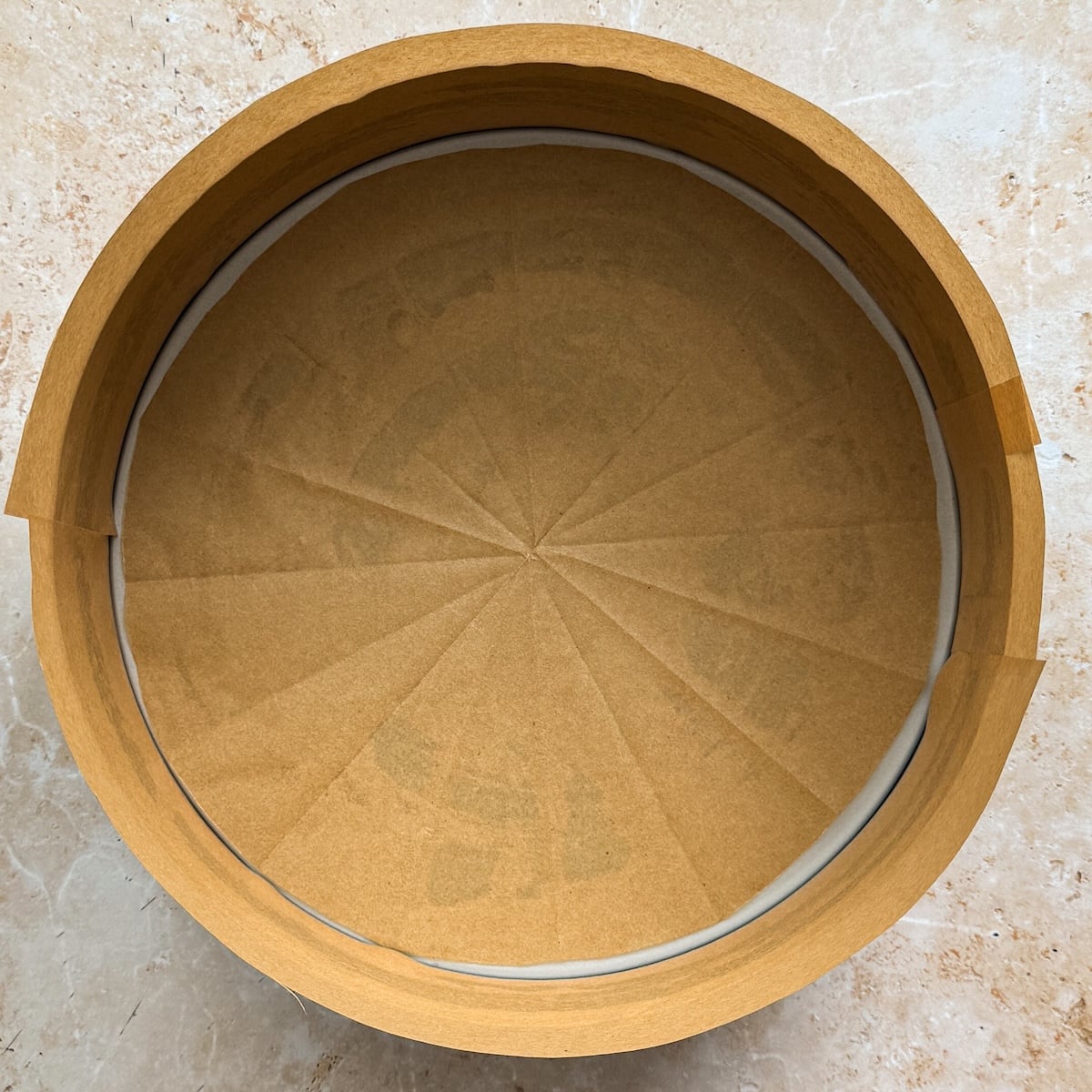 Round cake tin lined with parchment paper, cheesecake-ready with the paper neatly creased and folded along the sides and base to fit the tin. The tin sits on a light-colored, marbled surface.
