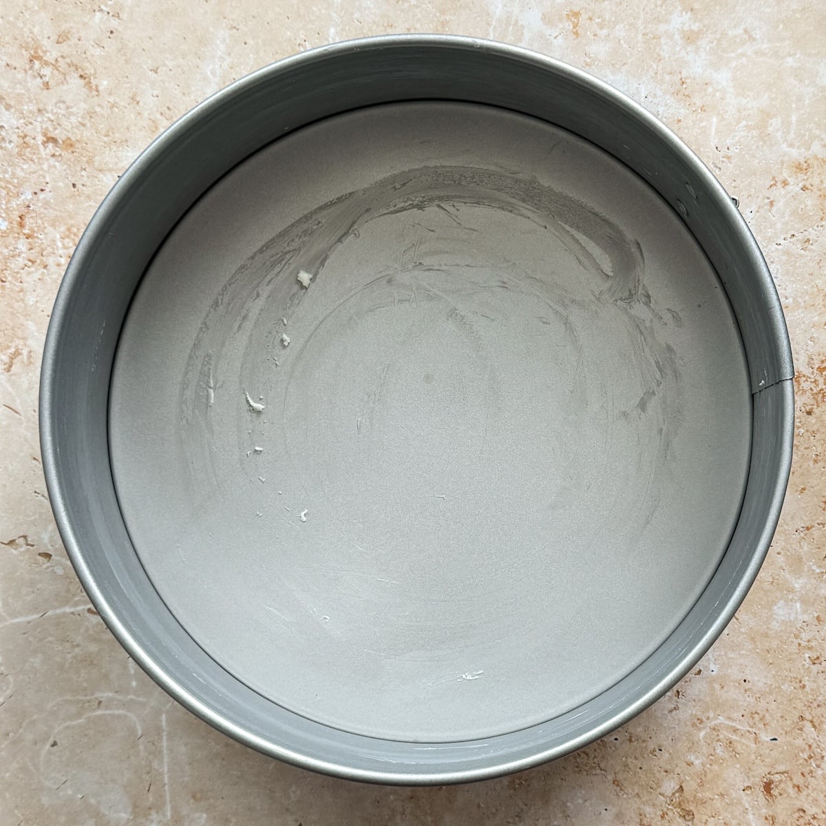 A round metal cake pan, perfect for baking cheesecake, with its interior coated in a thin layer of butter, sits on a light marbled countertop.