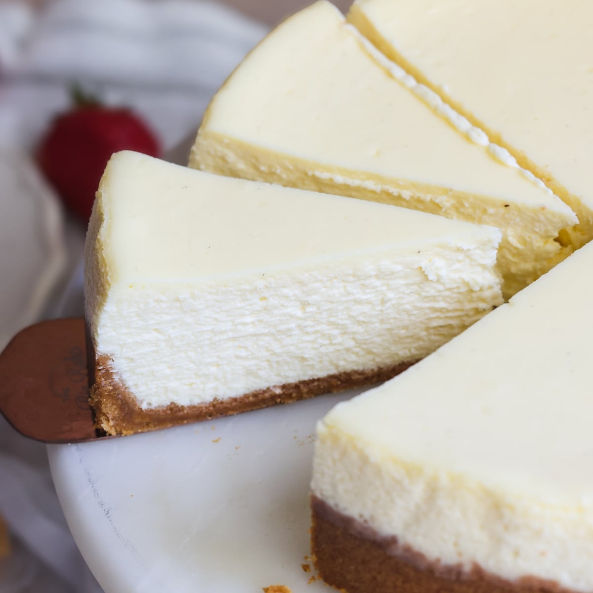 A slice of creamy cheesecake with a golden-brown graham cracker crust is being lifted from a cheesecake on a marble platter. It is smooth and crack free with a silky texture inside