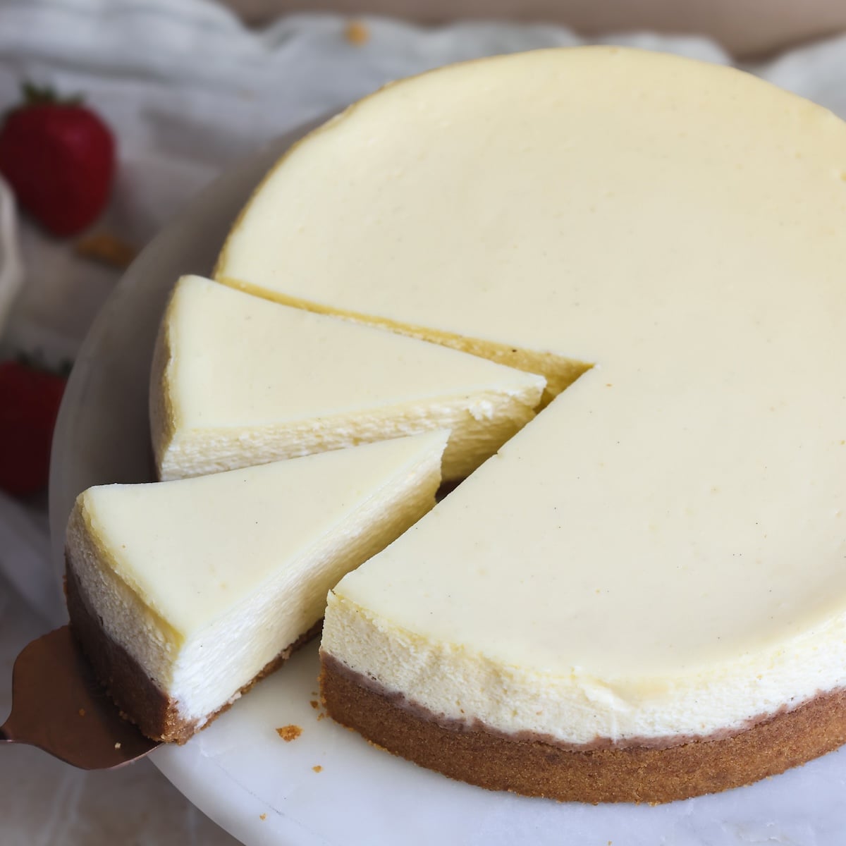 A classic cheesecake with a golden-brown crust sits on a white plate. One slice of rich cheesecake has been cut and is being lifted, showing the creamy texture inside. Fresh strawberries are visible in the background.