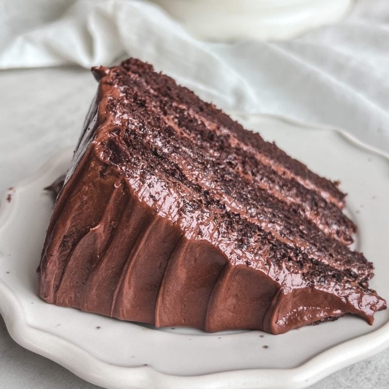 slice of chocolate cake on a plate revealing 4 moist layers with shiny cream cheese frosting