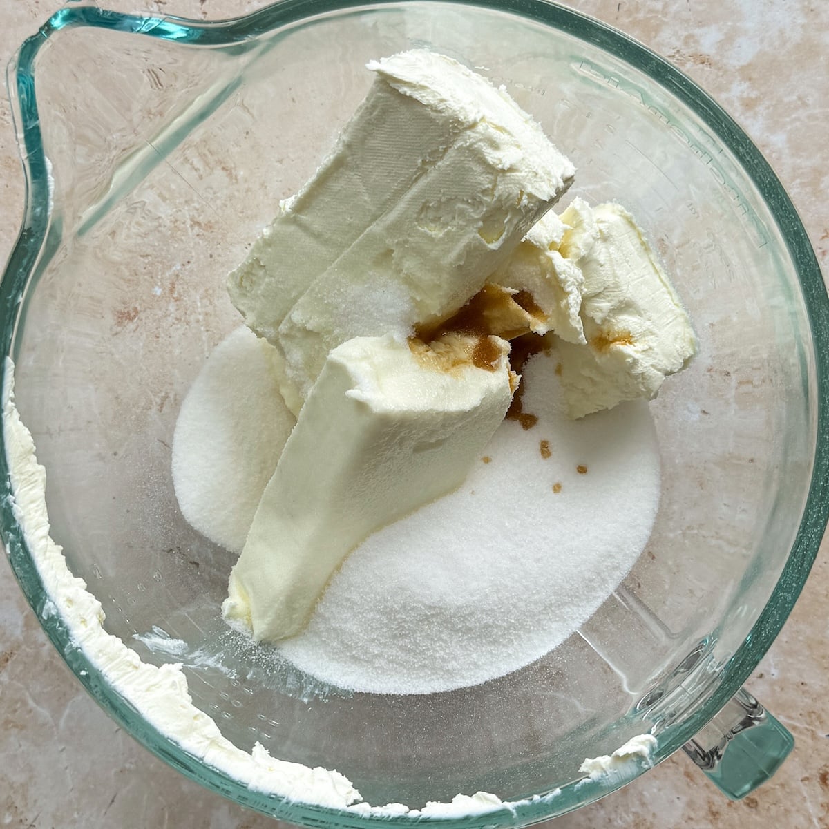 A glass mixing bowl with cream cheese, granulated sugar, and vanilla extract on a light countertop