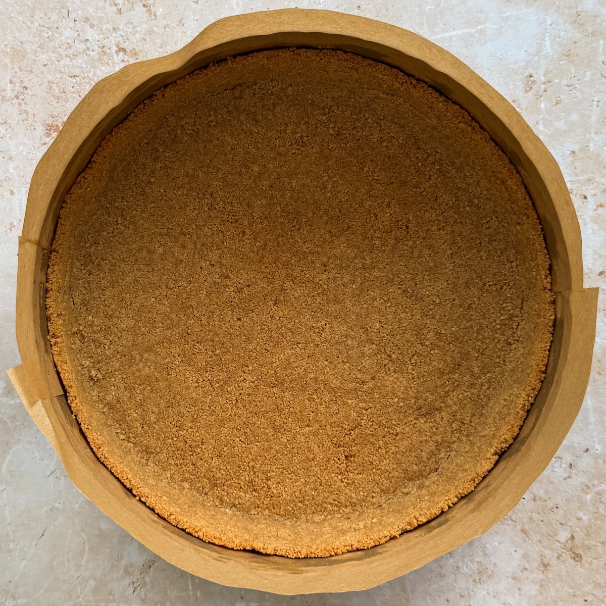 A round graham cracker pie crust sits in a brown paper-lined tin, viewed from above on a light-colored countertop.