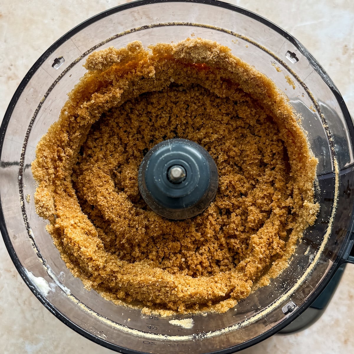 A food processor bowl filled with a coarse, crumbly brown crust mixture viewed from above. The mixture is spread evenly around the sides, leaving a circular space in the center where the blade attachment is visible.