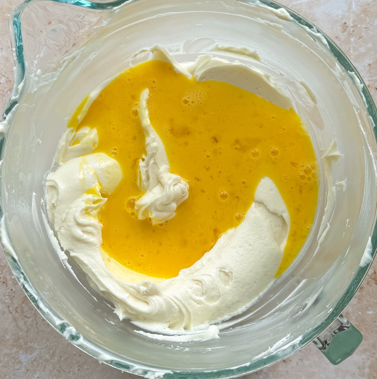 A glass mixing bowl holds a white cheesecake cream cheese mixture with yellow beaten eggs on top, ready to be combined, on a light countertop.