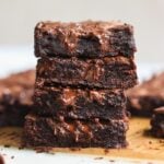 A close-up of four gooey, fudgy dark chocolate brownies stacked on top of each other, with melted chocolate visible on their surface. More brownies are blurred in the background on a wooden board.