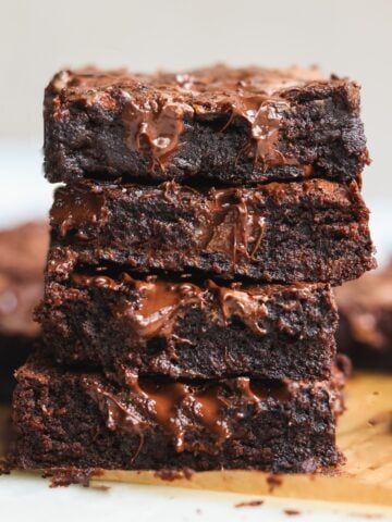 A close-up of four gooey, fudgy dark chocolate brownies stacked on top of each other, with melted chocolate visible on their surface. More brownies are blurred in the background on a wooden board.