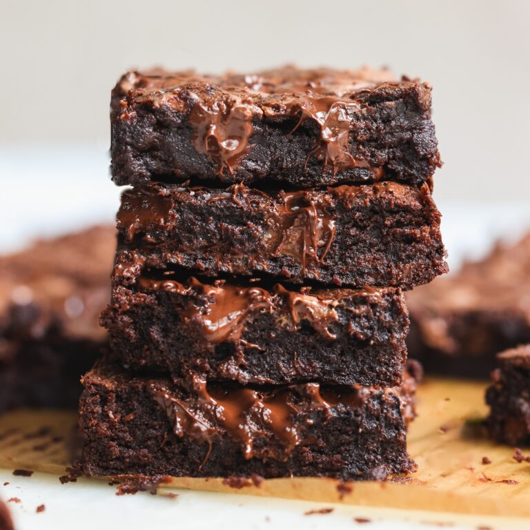 A close-up of four gooey, fudgy dark chocolate brownies stacked on top of each other, with melted chocolate visible on their surface. More brownies are blurred in the background on a wooden board.