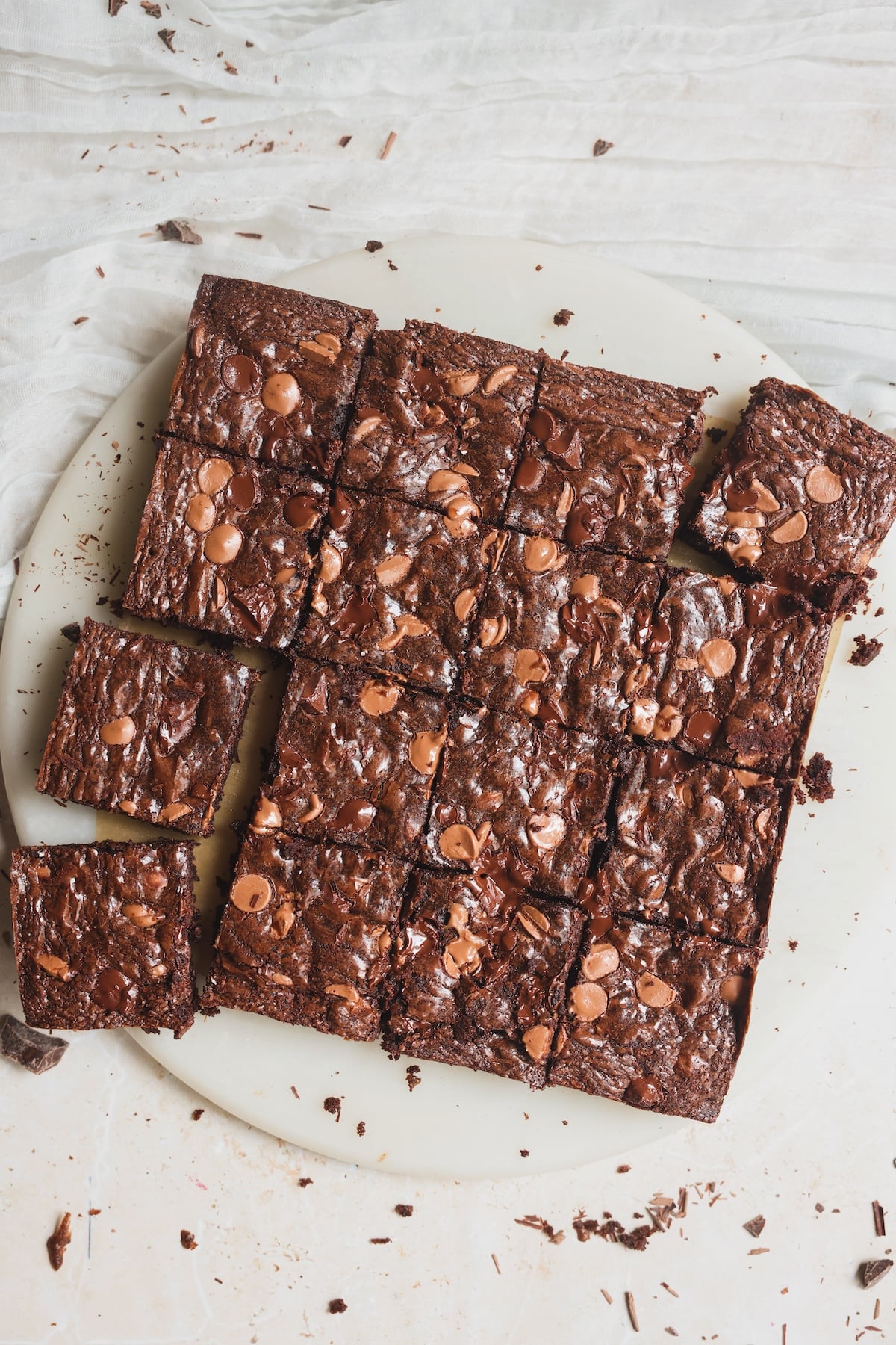 A round white plate with square-cut, best fudgy brownies topped with chocolate chips, arranged in a neat grid on a light textured background. Some crumbs are scattered around the plate.