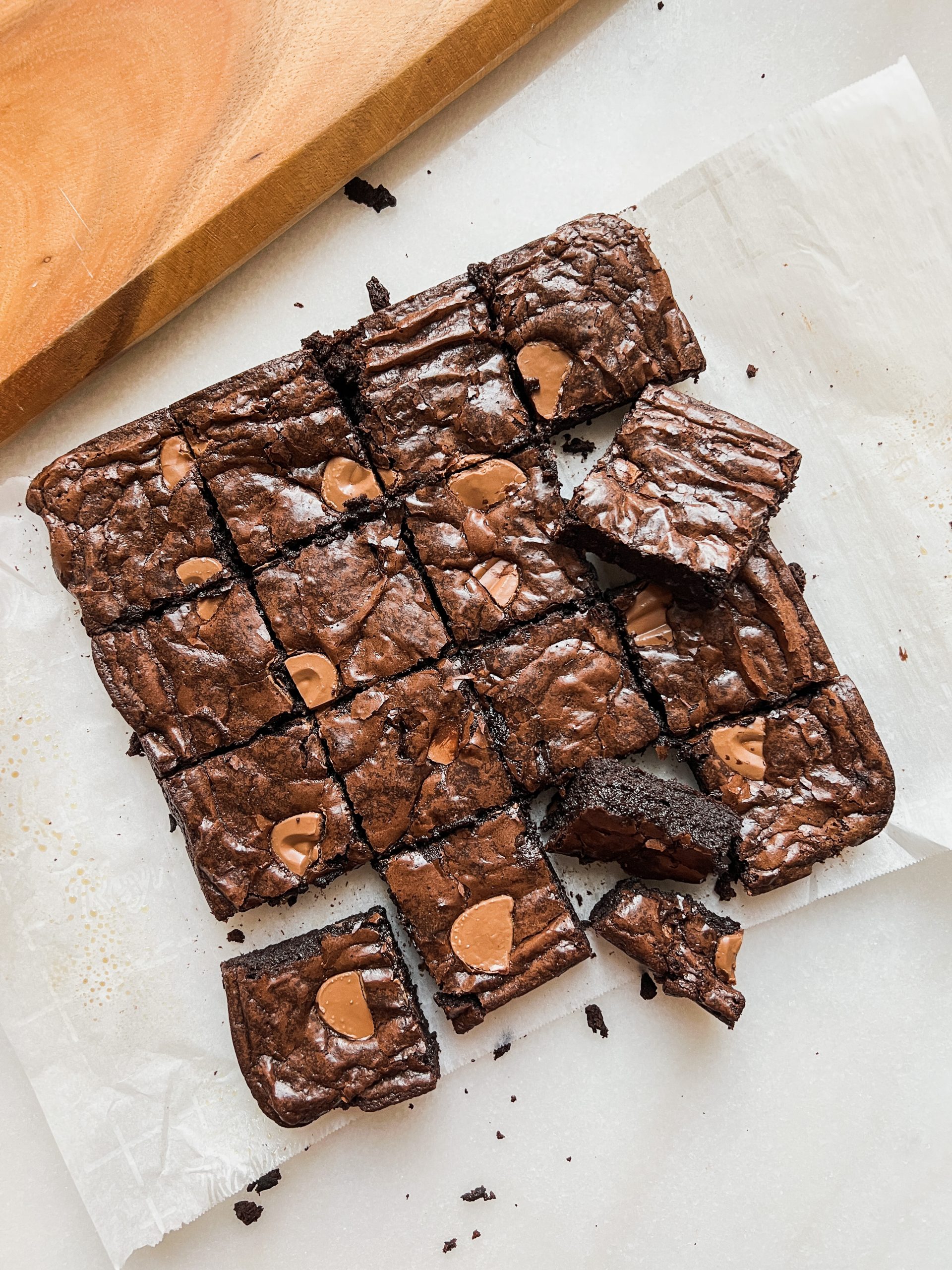 Angled overhead shot of a pan of 16 brownies placed on parchment paper. Brownies have a dark fudgy interior and crinkly shiny top with large chocolate chunks