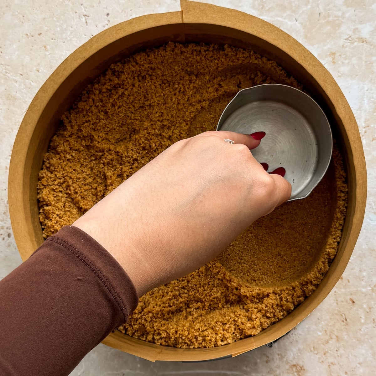 A hand presses down a crumbly brown cheesecake crust with a metal cup inside a round baking pan on a light countertop.