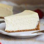 A slice of classic cheesecake with a golden-brown crust sits on a white plate, with a fork beside it and a blurred strawberry in the background, highlighting the creamy richness of this delicious cheesecake dessert.