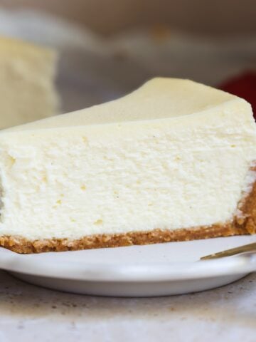 A slice of classic cheesecake with a golden-brown crust sits on a white plate, with a fork beside it and a blurred strawberry in the background, highlighting the creamy richness of this delicious cheesecake dessert.