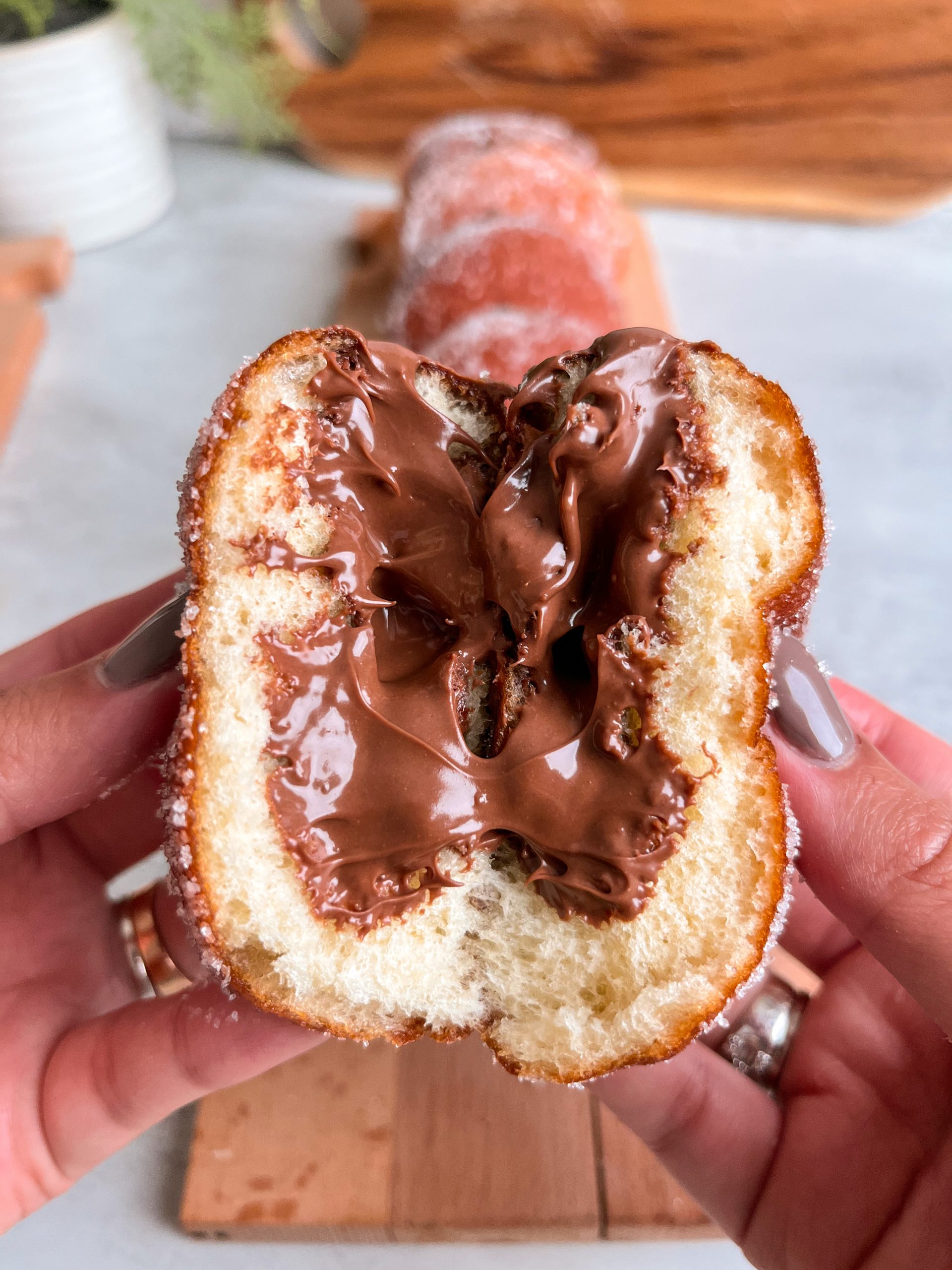 A halved donut held in two hands. Filled with nutella oozing out of the center. Donut has a soft, fine crumb.