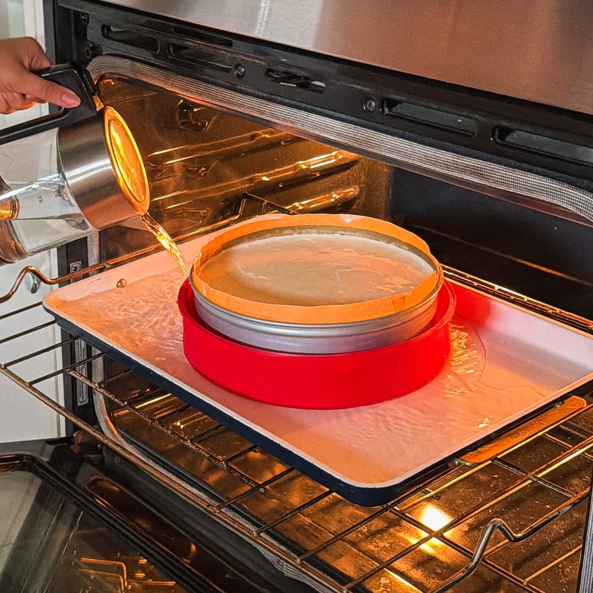 A person pours hot water into a baking tray in an oven, surrounding a cheesecake pan with a red silicone wrap, carefully preparing a water bath for cheesecake.