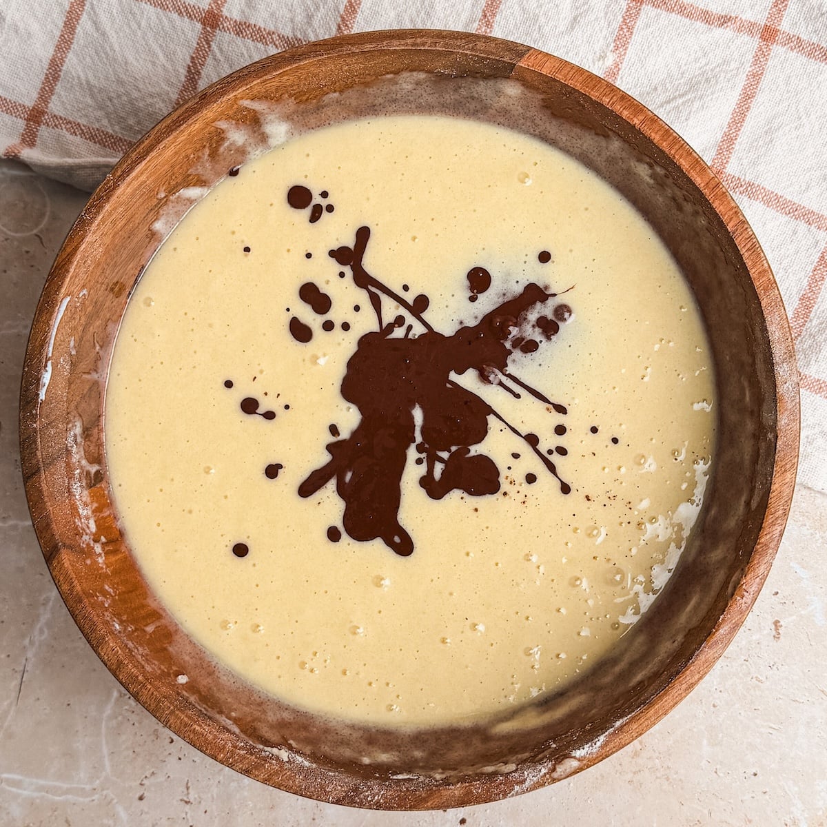 A wooden bowl filled with smooth yellow cake batter, topped with a splash of cocoa powder mixed with water. A beige towel with a checkered pattern is partially visible in the background.