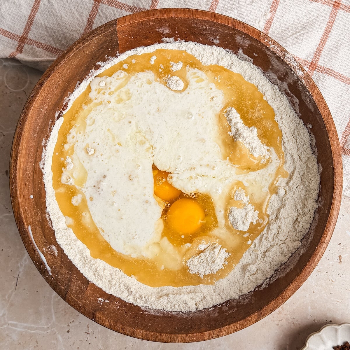 A wooden bowl with flour, cracked eggs, and buttermilk being mixed for a chocolate cake recipe, ready for baking, on a beige countertop beside a plaid kitchen towel.