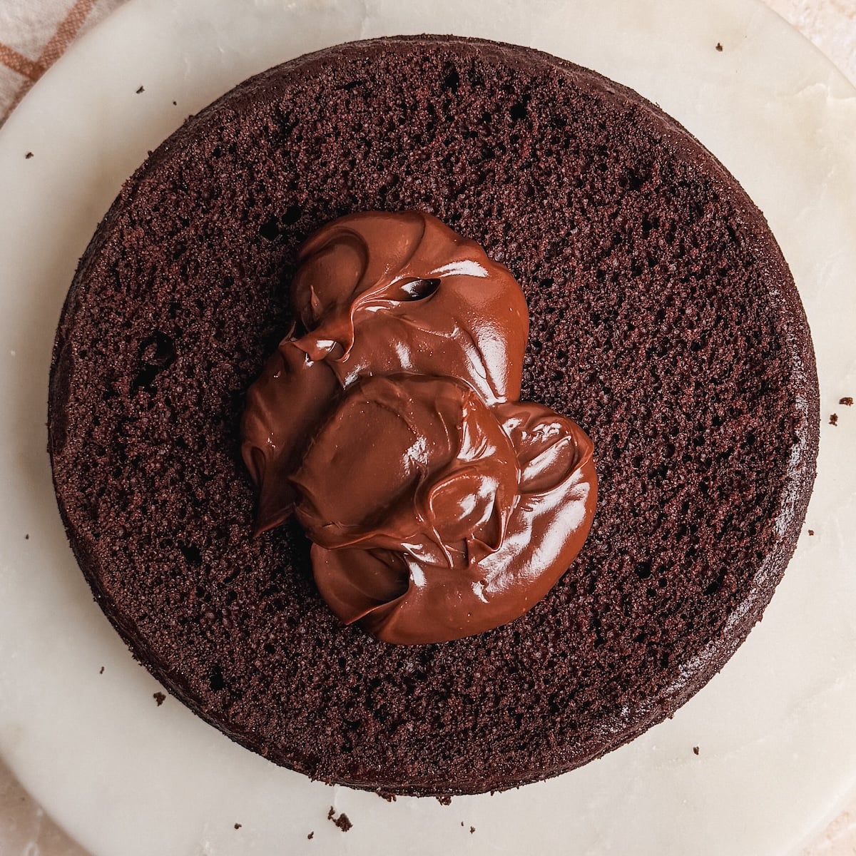 A round chocolate cake layer on a marble surface with a large dollop of chocolate ganache in the center, ready to be spread