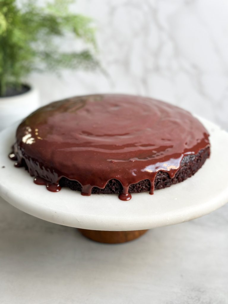 Round chocolate cake on a marble serving stand with a shiny chocolate ganache on the top dripping from the sides