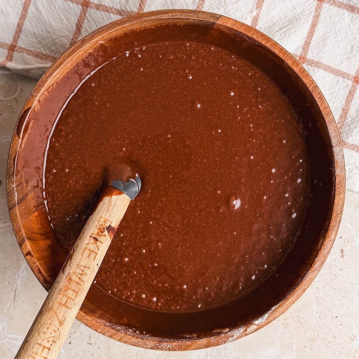 A wooden bowl filled with chocolate cake recipe batter, with a spatula resting in it. A beige and white checkered cloth is partially visible in the background.