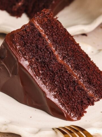 A slice of rich chocolate cake with two layers and smooth chocolate ganache frosting served on a white plate with a gold fork beside it.