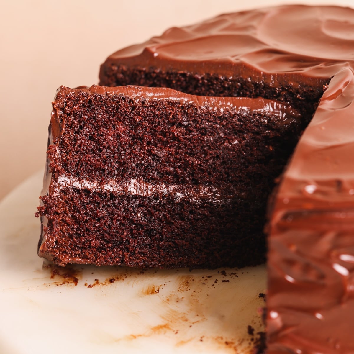 A close-up of a rich, moist chocolate cake with thick chocolate frosting. One large slice is being pulled out, revealing the cake’s dark chocolate layers and creamy filling. The cake sits on a white plate.