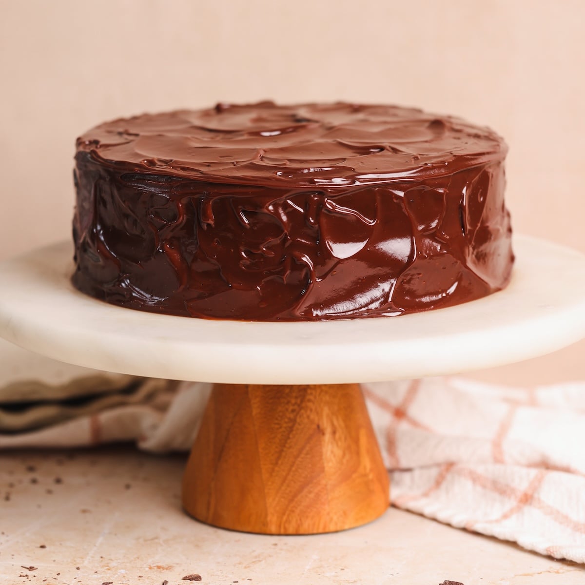 A round chocolate cake with a glossy chocolate ganache frosting sits on a marble cake stand with a wooden base. A beige cloth with a checkered pattern is in the background.