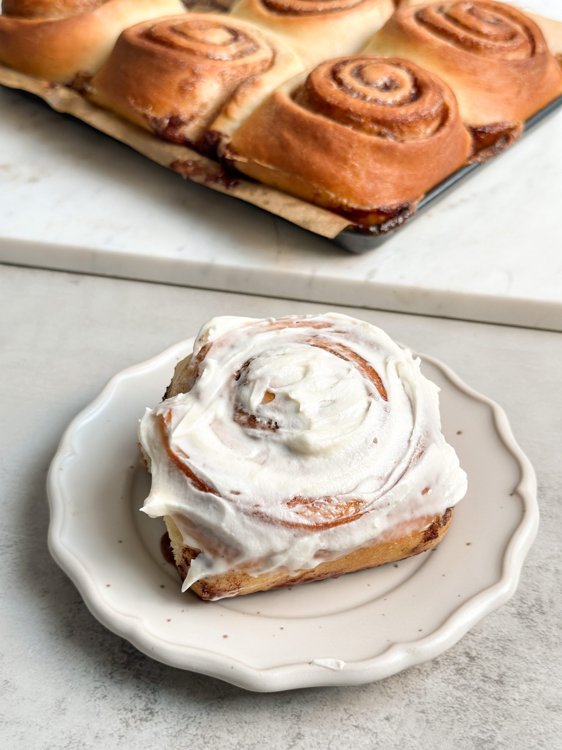 A fluffy cinnamon roll on a small plate, with cream cheese frosting on it. Batch of cinnamon rolls seen in background