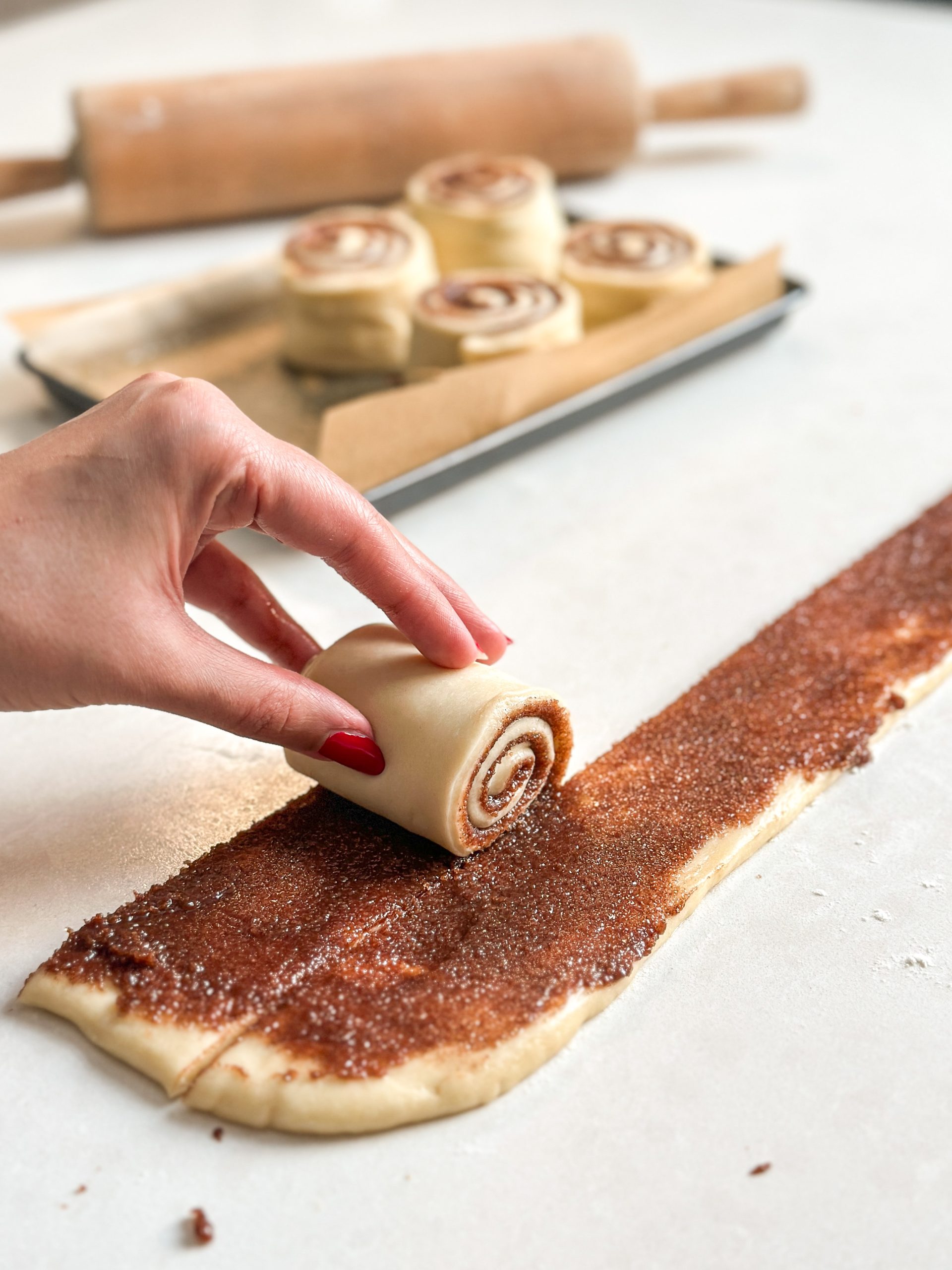 Hand is rolling a strip of dough with cinnamon sugar on it into a neatly rolled cinnamon roll