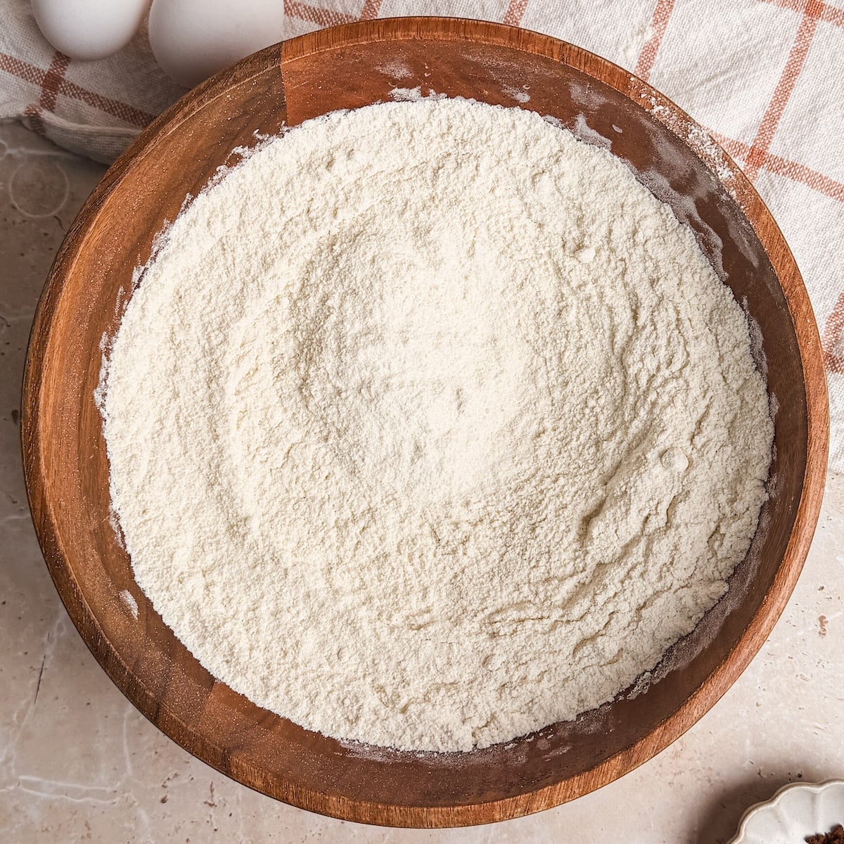 A wooden bowl filled with mixed dry ingredients for cake on a light countertop, near a beige and red plaid kitchen towel and two eggs in the background.