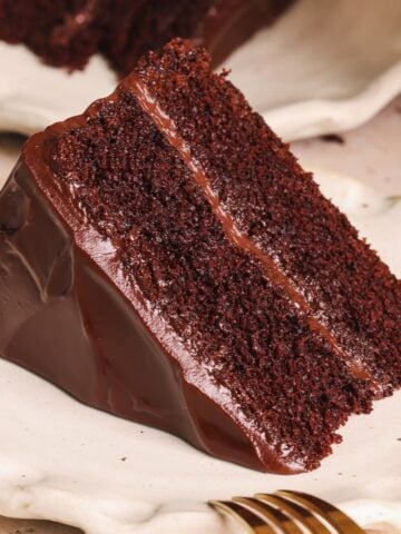 A slice of rich, moist chocolate cake made from an easy chocolate cake recipe, with a shiny chocolate ganache frosting between layers and on the outside, served on a white plate with a gold fork nearby.