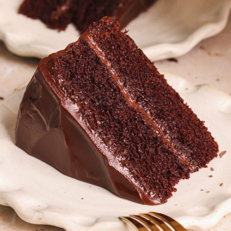A slice of rich, moist chocolate cake made from an easy chocolate cake recipe, with a shiny chocolate ganache frosting between layers and on the outside, served on a white plate with a gold fork nearby.