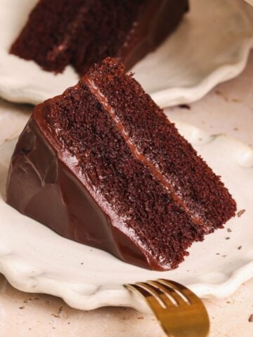 A slice of chocolate cake on a small plate with a fork resting on it, and another slice in the background. cake has 2 layers with a silky ganache frosting