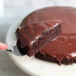 Slice of chocolate cake with shiny ganache on it with the rest of the cake in the background