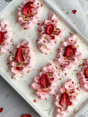 overhead picture of a dish of tres leches cake covered in whipped cream and piped pink swirls. it is sprinkled with freeze dried strawberries