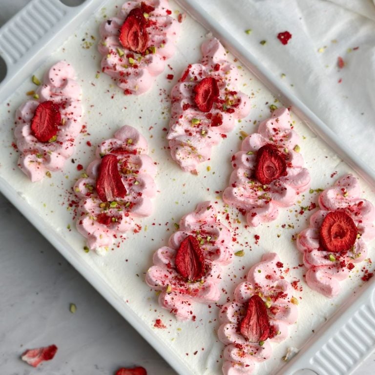overhead picture of a dish of tres leches cake covered in whipped cream and piped pink swirls. it is sprinkled with freeze dried strawberries