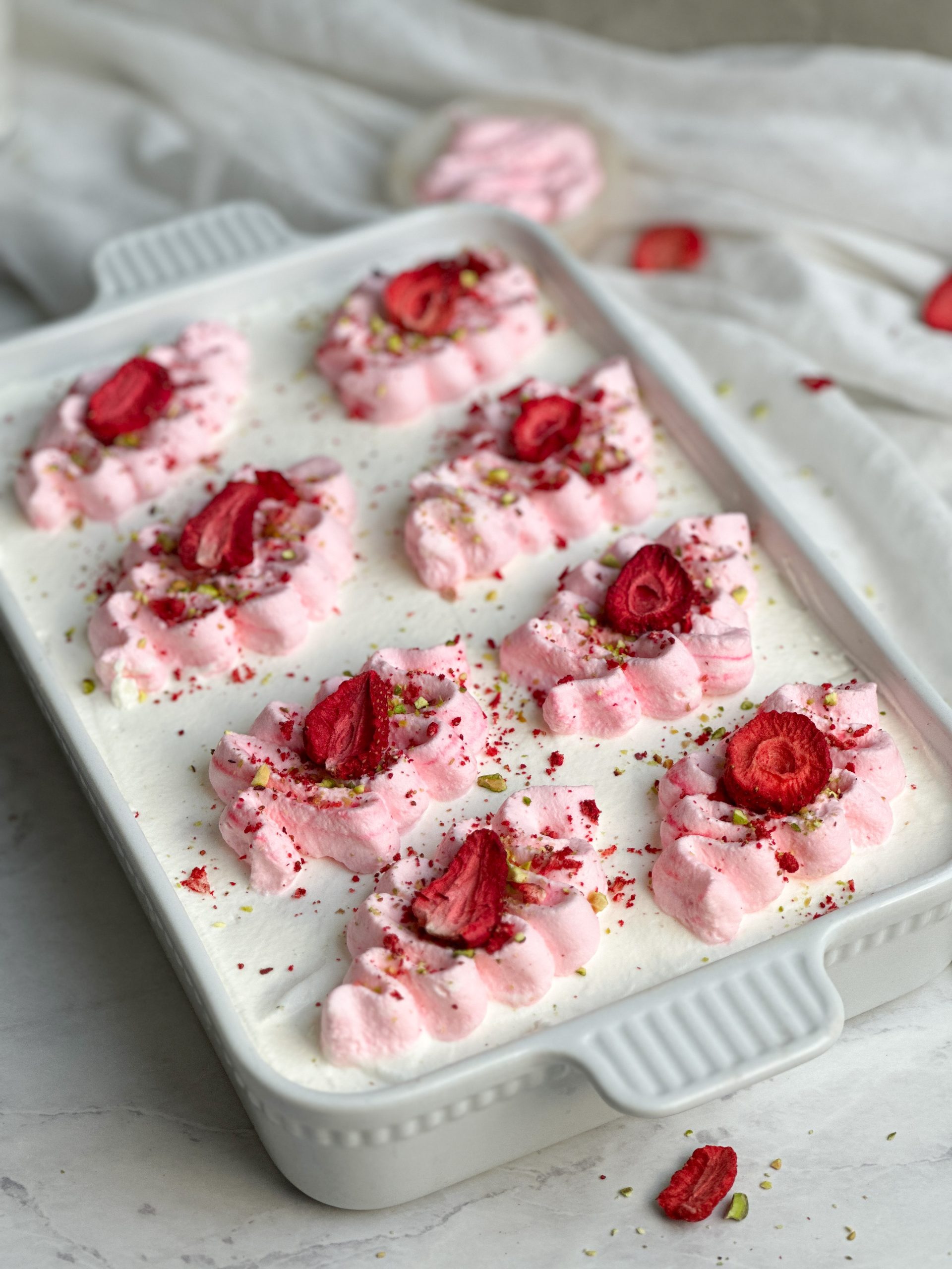 a baking dish with tres leches cake inside. cake has whipped cream spread on it, and light pink ribbons piped on top. it is decorated with freeze dried strawberries and chopped pistachios