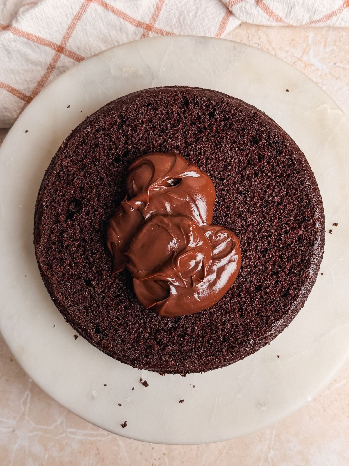 ganache frosting added on top of layer of chocolate cake on a marble cake stand