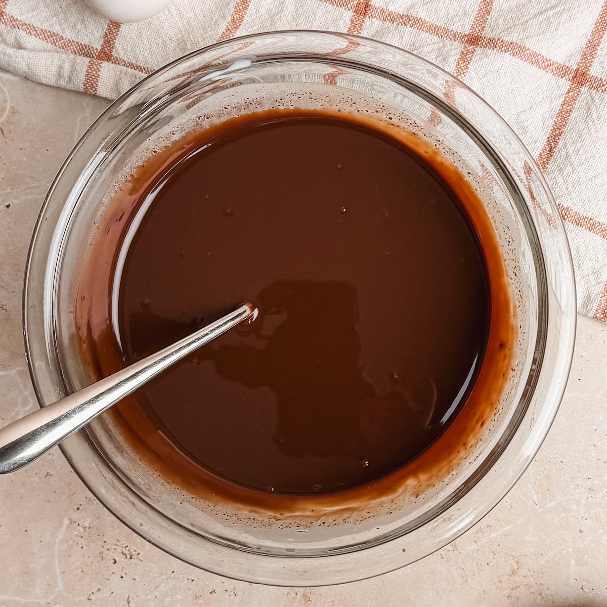 A glass bowl filled with smooth, melted chocolate ganache and a metal spoon inside, set on a light countertop with a beige and orange plaid cloth nearby.