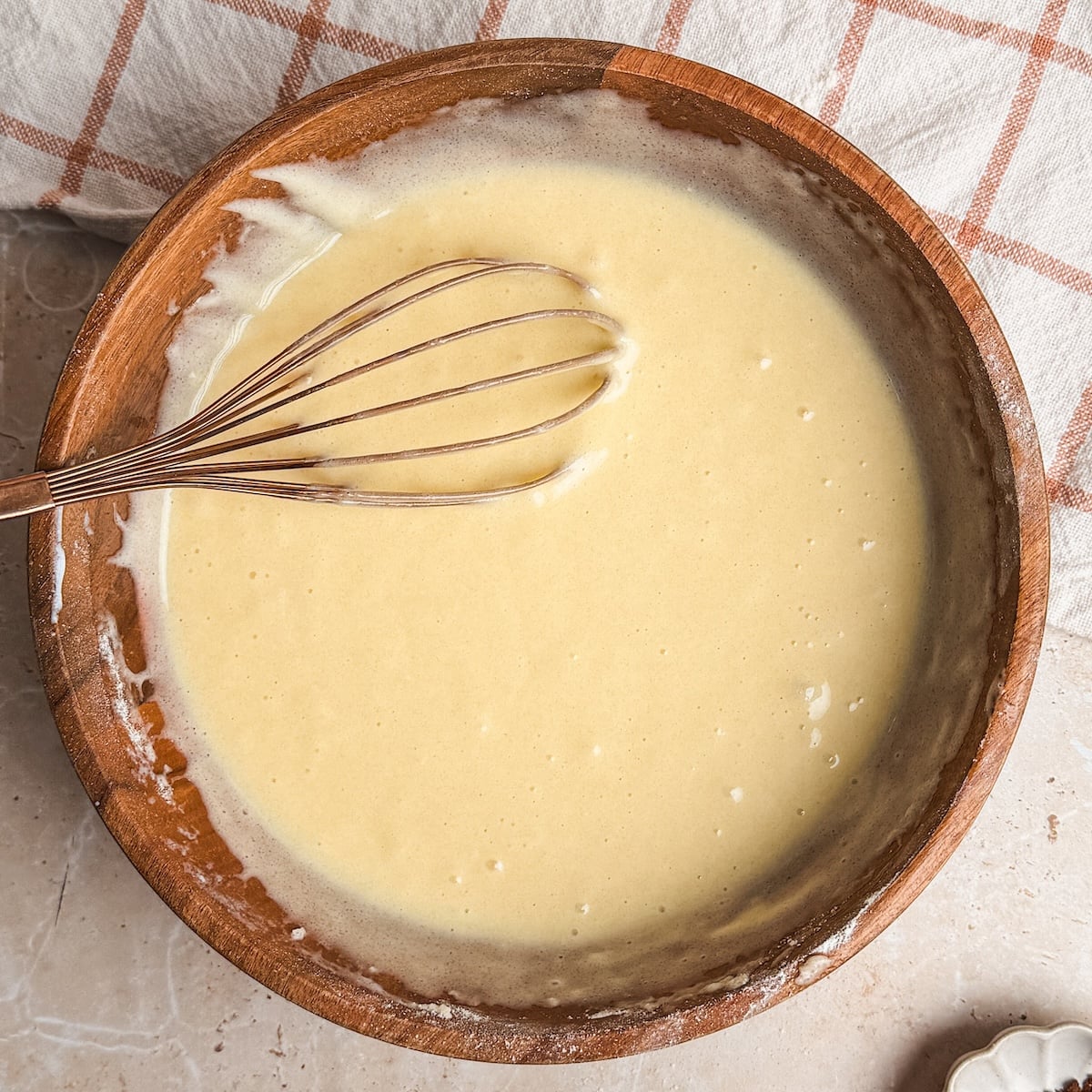 A wooden bowl filled with smooth cake batter and a metal whisk rests on a counter next to a beige and orange checkered cloth