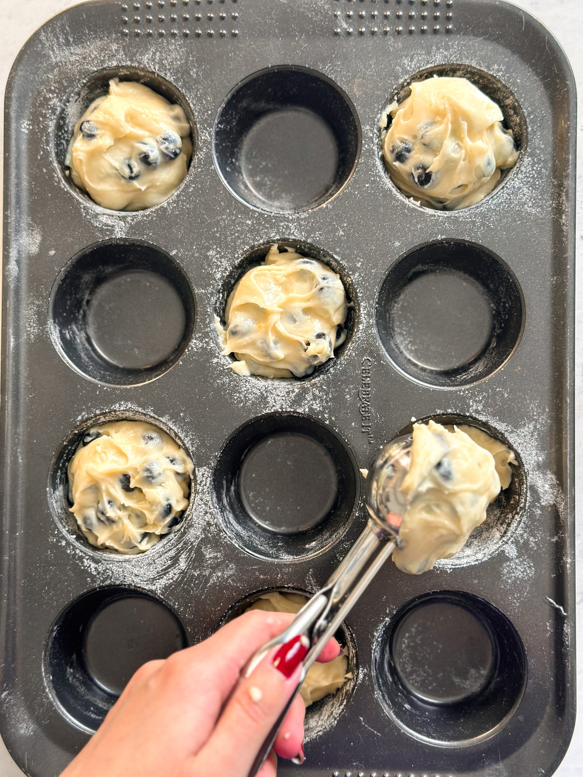 overhead picture of a hand scooping blueberry muffin batter into a muffin tray