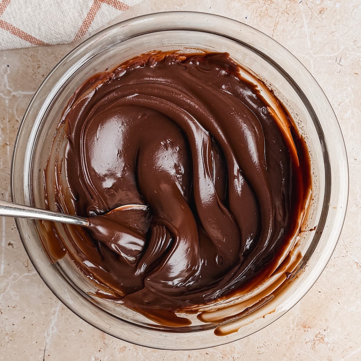A glass bowl filled with smooth, glossy chocolate ganache being stirred with a metal spoon on a light marble surface. It looks thick and spreadable