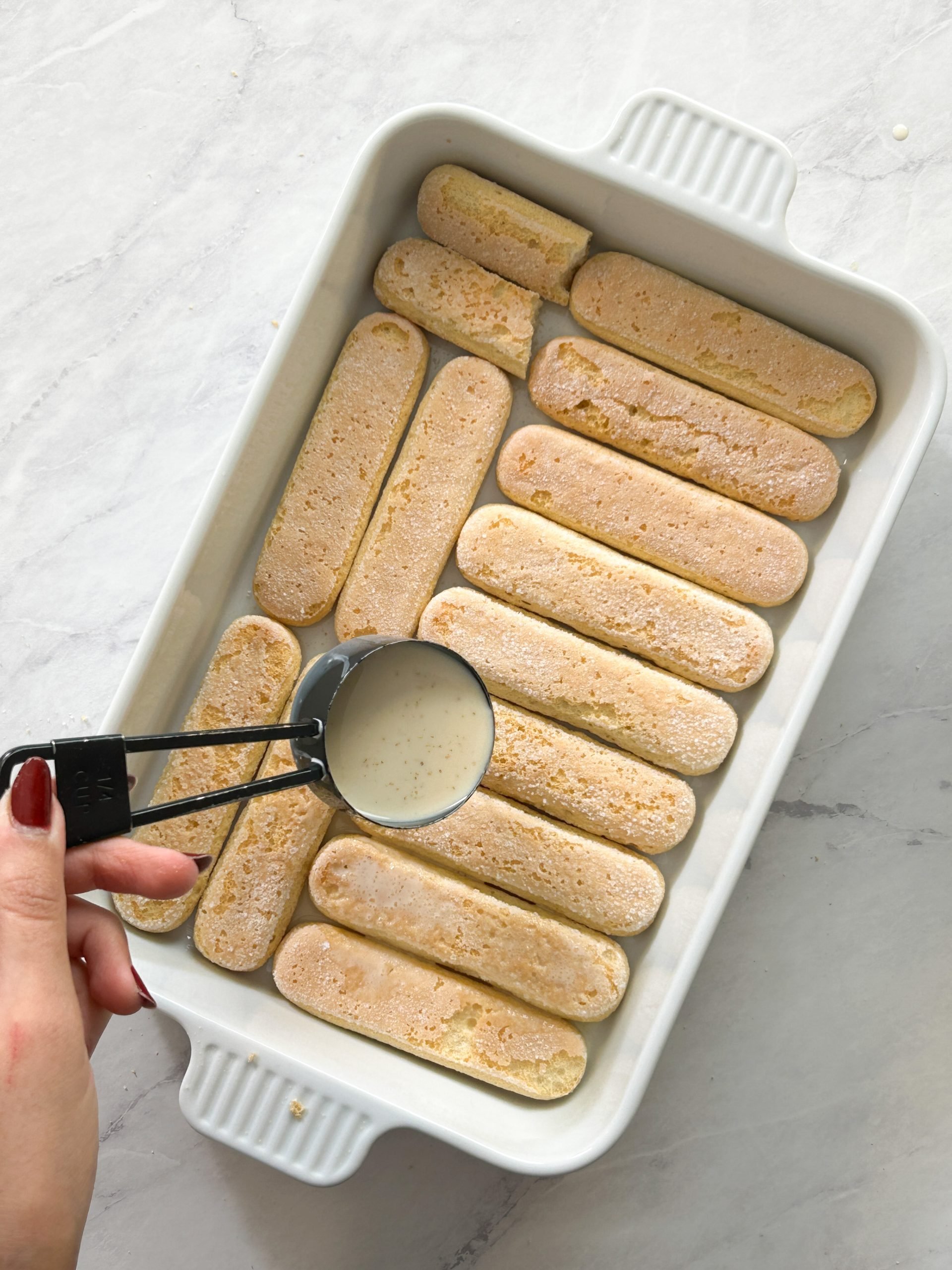 a hand is pouring tres leches milk mixture onto the ladyfingers in a dish