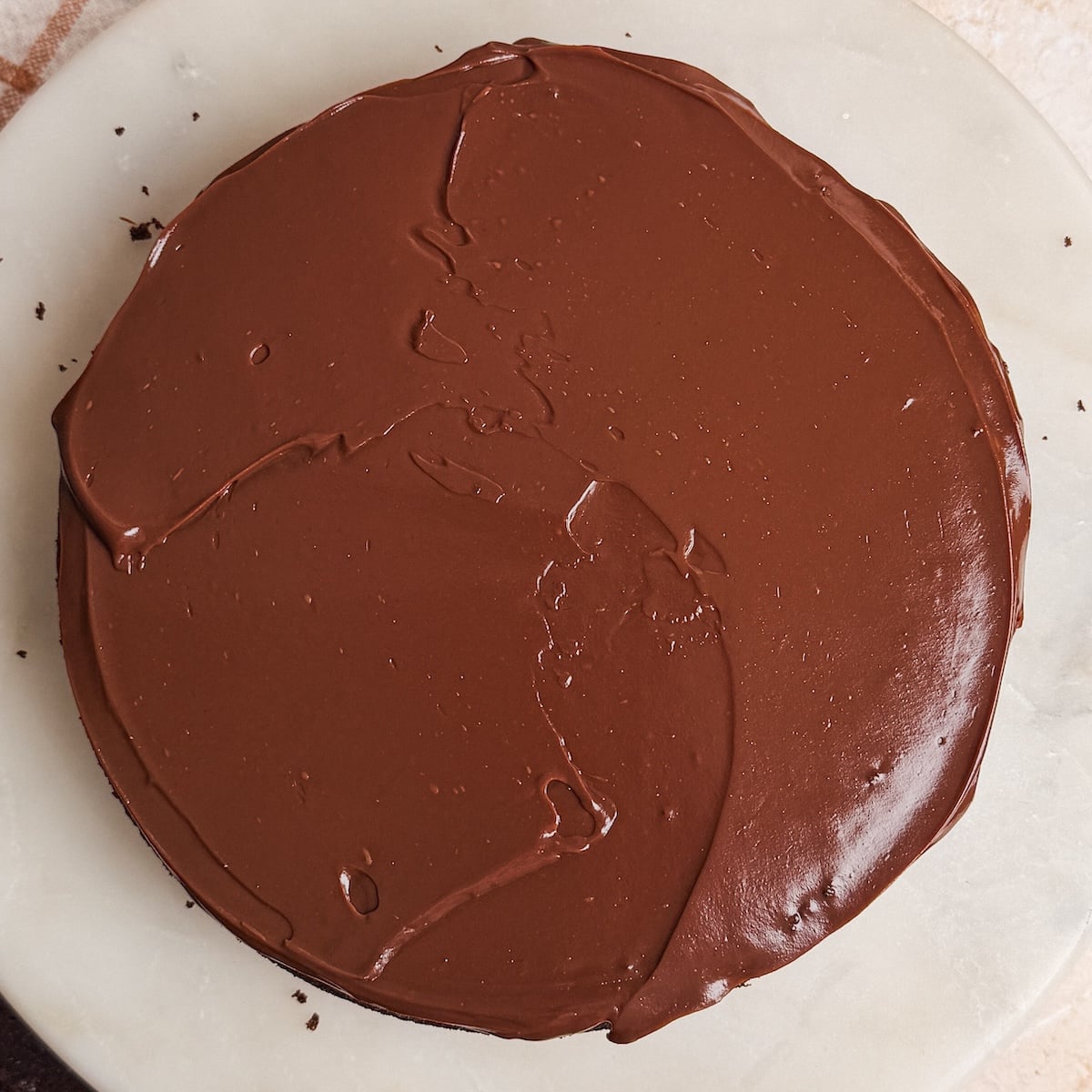 A round chocolate cake covered smoothly with glossy chocolate ganache, sits on a white marble surface.