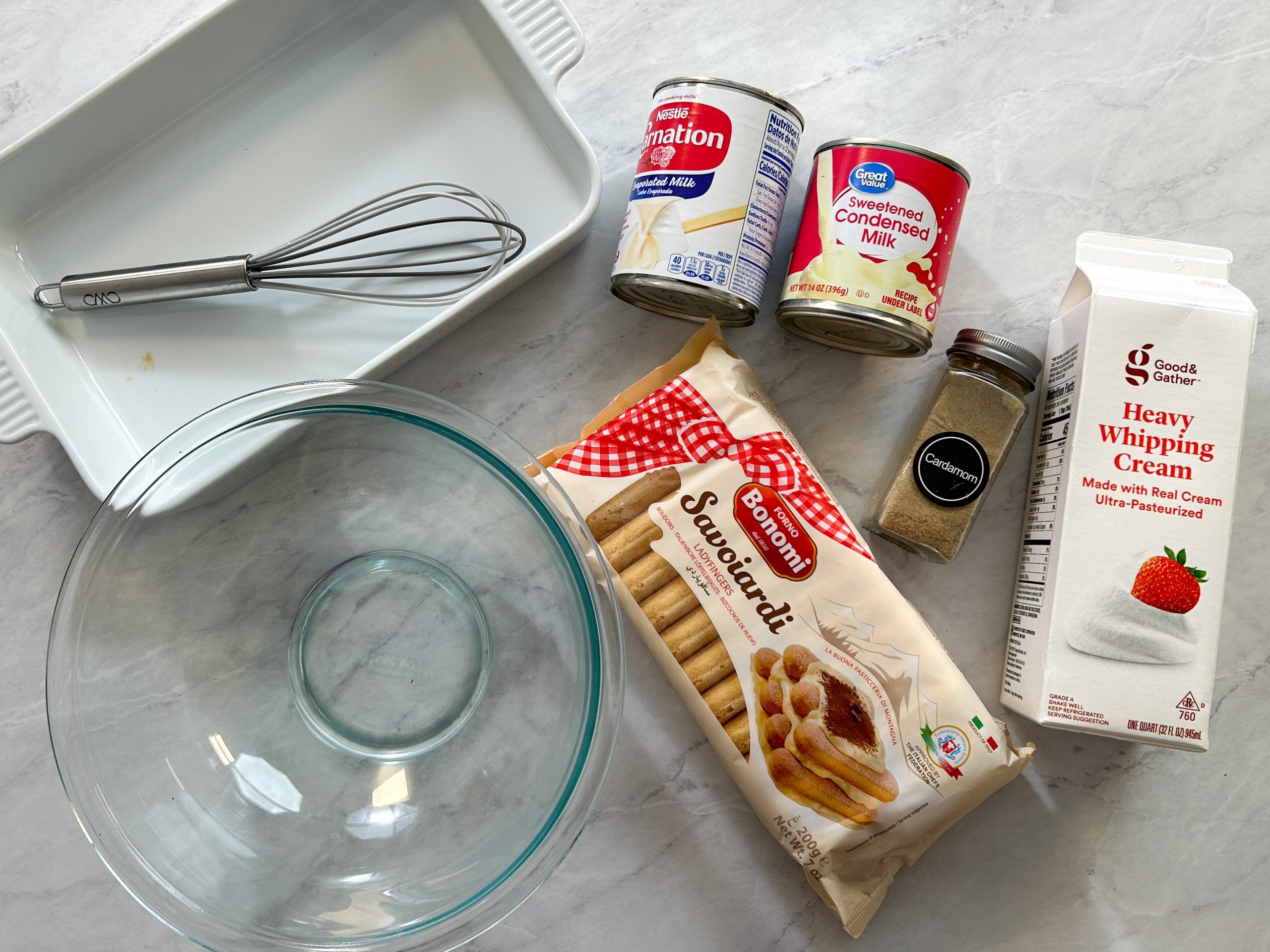 overhead picture of all ingredients required to make tres leches cake: bowl, pan, whisk, ladyfingers, cream, sweetened condensed milk, evaporated milk, and cardamom